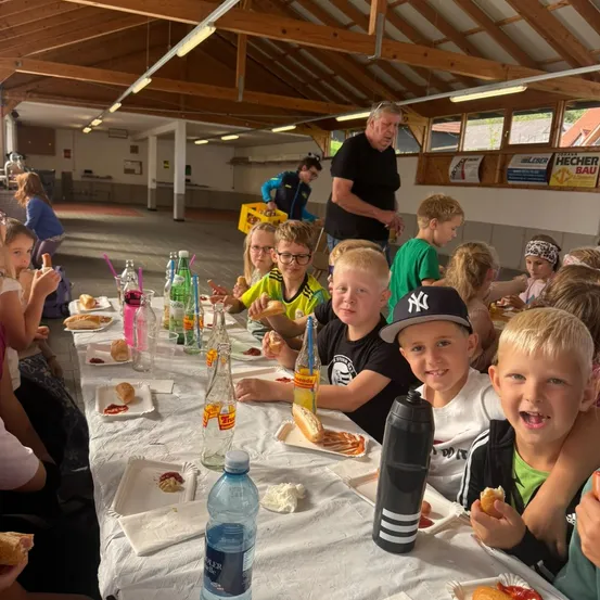 Eine Gruppe von Kindern und Erwachsenen sitzt an einem langen Tisch in einem Saal und isst Essen und Getränke.