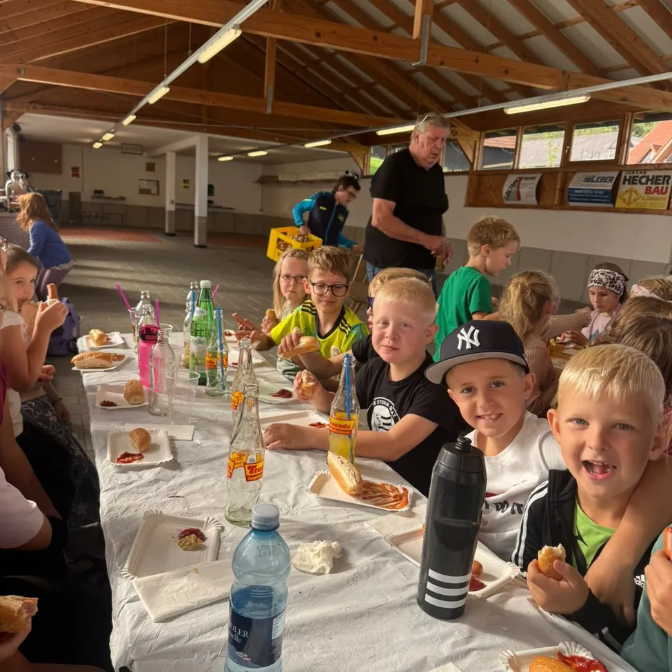 Eine Gruppe von Kindern und Erwachsenen sitzt an einem langen Tisch in einem Saal und isst Essen und Getränke.