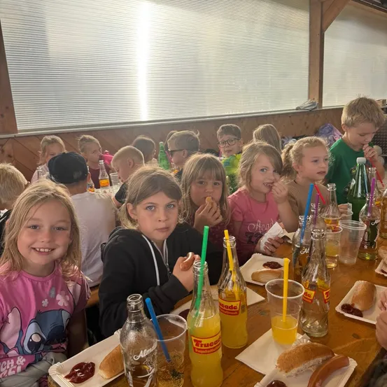 Eine Gruppe von Kindern sitzt an einem langen Holztisch und isst Hot Dogs und trinkt Orangensaft.