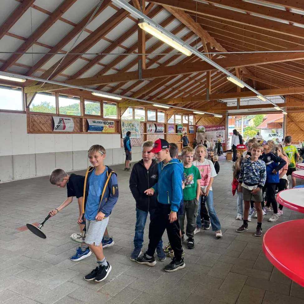 Eine Gruppe von Kindern versammelt sich in einem überdachten Bereich, einige spielen mit einer Schaufel, während andere stehen und zuschauen.