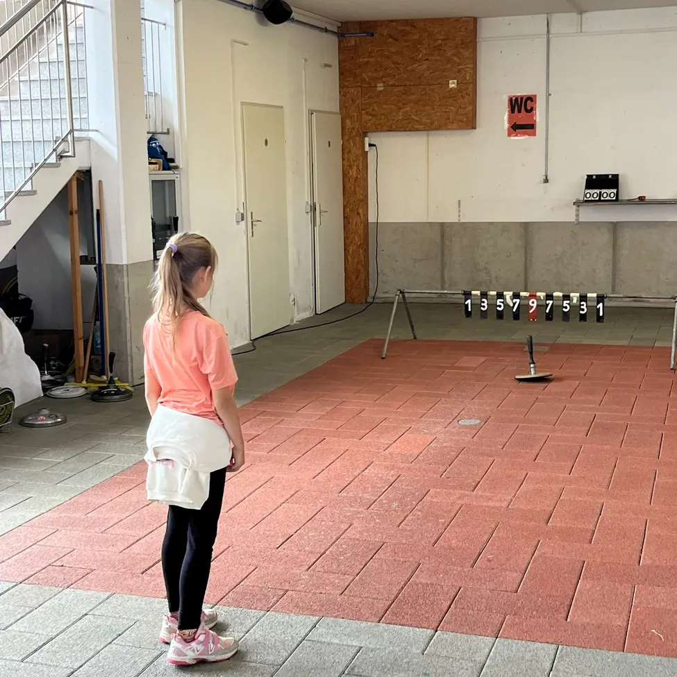 Ein Mädchen in einem rosa Shirt und schwarzen Leggings steht in einem Raum mit einem roten Boden und schaut auf eine Bowlingbahn.