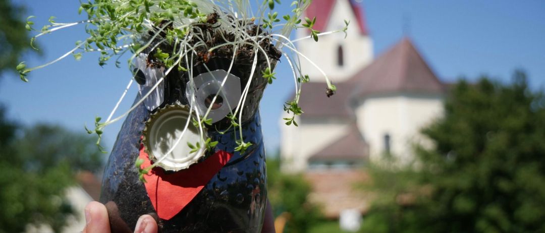 Ein kreatives Handwerk mit einer blauen Flaschenvasen in Form einer Eule, aus der Pflanzen sprießen, und einem Deckel, der als Auge dient, vor einer Kirche gehalten.
