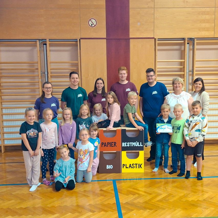 Eine Gruppe von Kindern und Erwachsenen steht zusammen in einer Turnhalle, einige sitzen auf Recycling-Behältern. Sie alle lächeln für die Kamera.