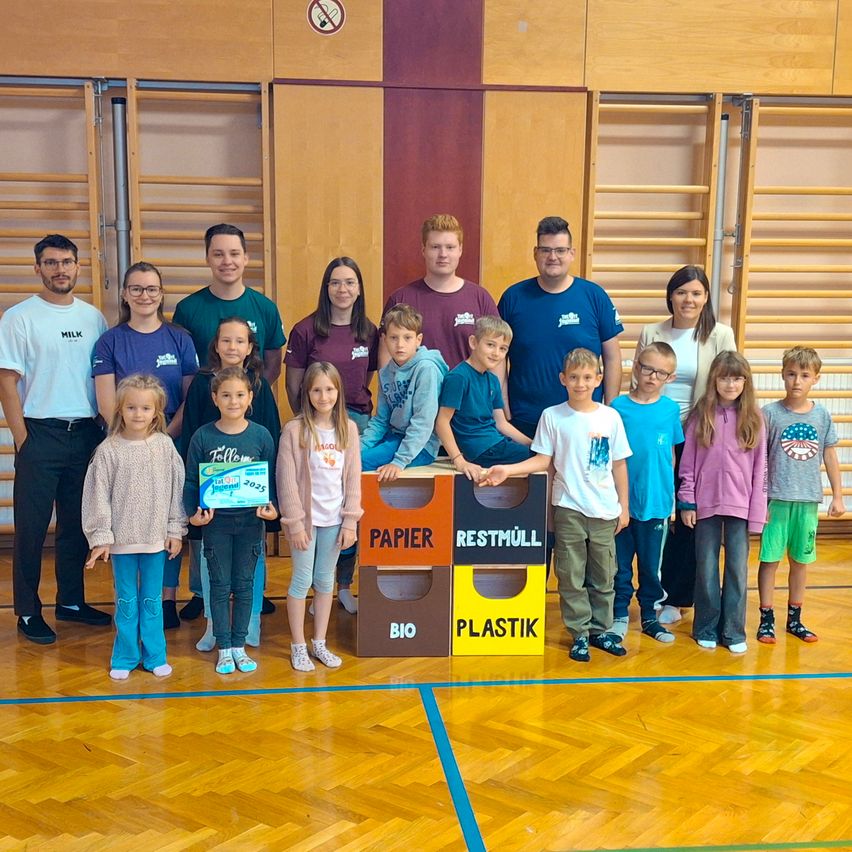 Gruppenfoto mit Recycling-Behältern: Erwachsene und Kinder in Freizeitkleidung, stehend und sitzend, einige halten Auszeichnungen, in einem Raum mit Holzwänden und -boden.