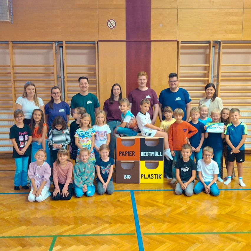 Eine Gruppe von Kindern und Erwachsenen posiert für ein Foto in einer Turnhalle, in der Nähe von Recycling-Behältern mit den Aufschriften Papier, Restmüll, Bio und Plastik.