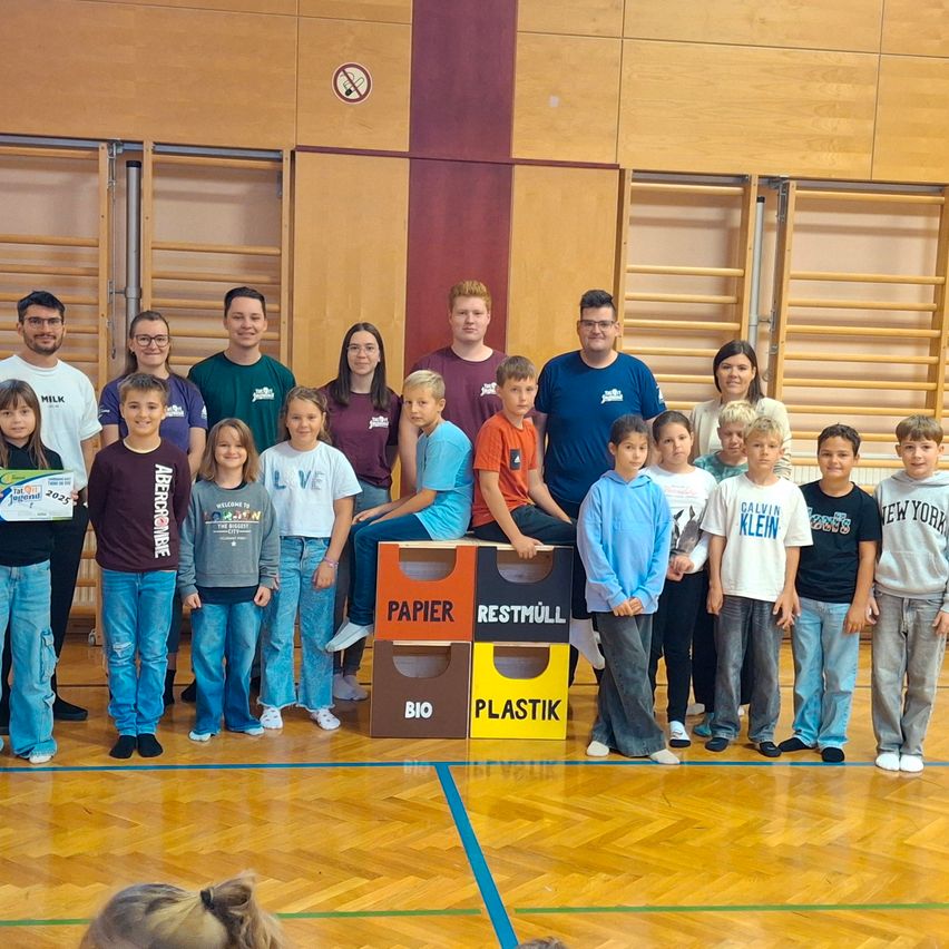 Eine Gruppe von Menschen, einschließlich Kindern und Erwachsenen, posiert für ein Foto in einer Turnhalle mit Recycling-Behältern vor ihnen.