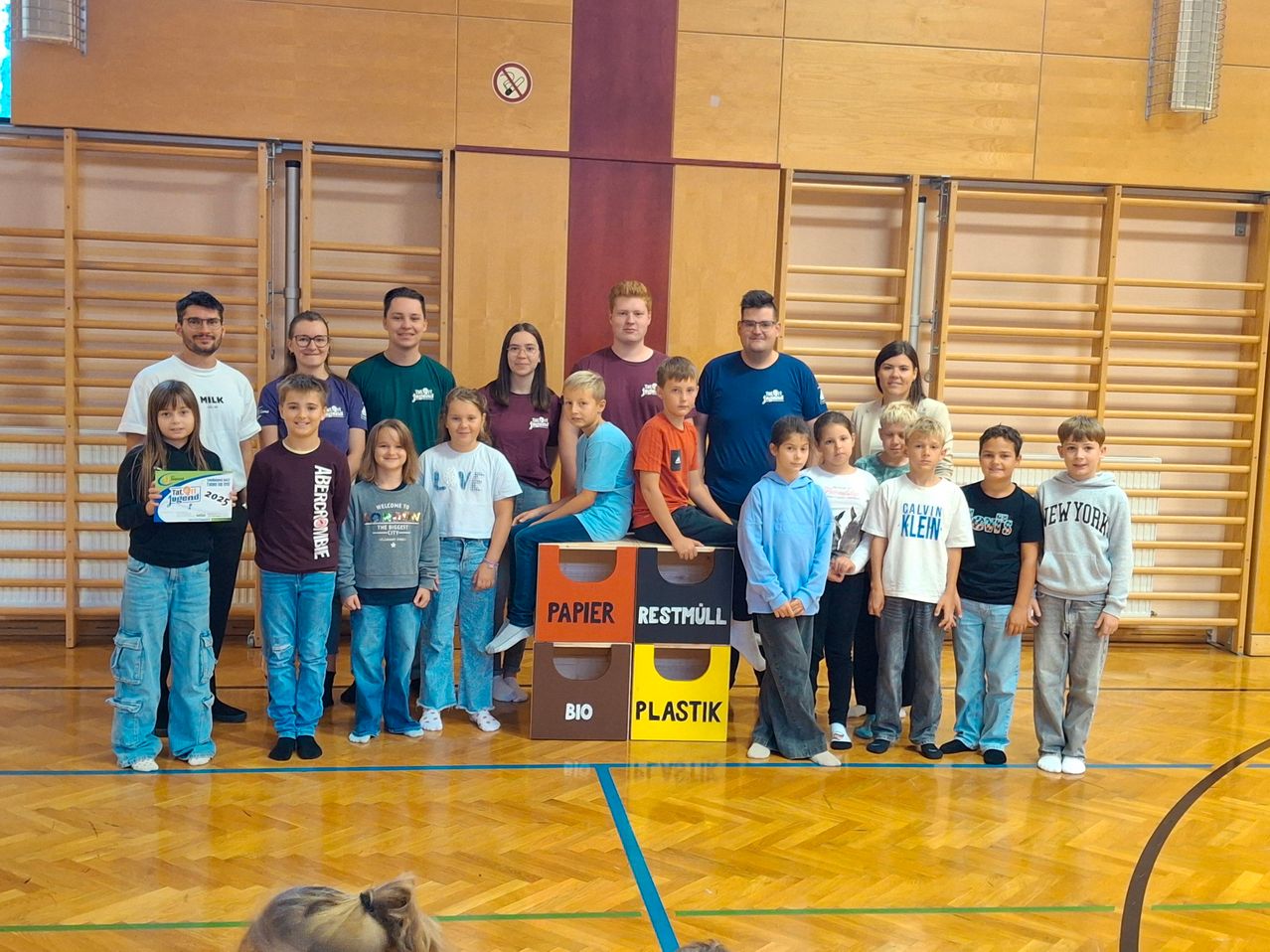 Eine Gruppe von Menschen, einschließlich Kindern und Erwachsenen, posiert für ein Foto in einer Turnhalle mit Recycling-Behältern vor ihnen.