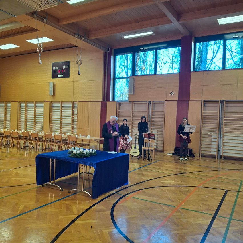 Ein älterer Mann steht in einer Turnhalle mit einem in Blau gedeckten Tisch. Vier Frauen, zwei mit Gitarren, stehen hinter ihm. Der Raum hat Holzböden, Wände und Fenster.