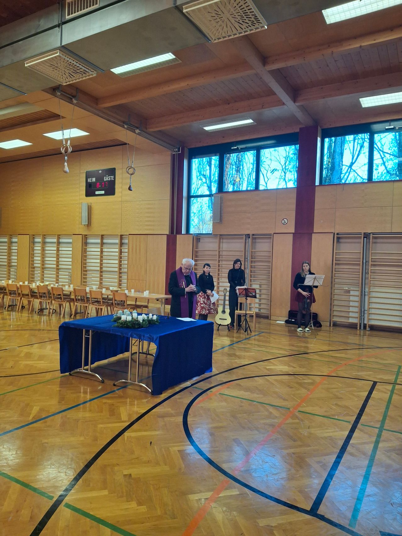 Ein älterer Mann steht in einer Turnhalle mit einem in Blau gedeckten Tisch. Vier Frauen, zwei mit Gitarren, stehen hinter ihm. Der Raum hat Holzböden, Wände und Fenster.