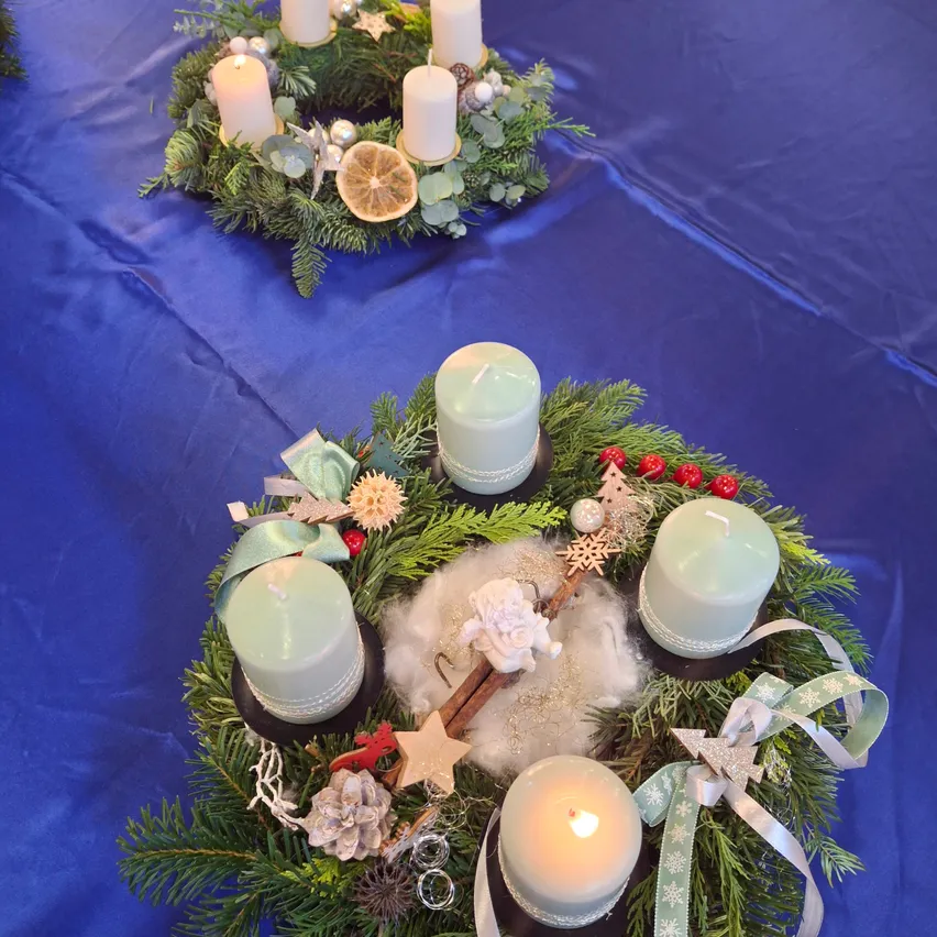 Zwei Kränze mit Kerzen, Tannenzapfen, Schleifen und Ornamenten ruhen auf einem blauen Tischtuch. Ein Kranz hat eine Zitronenscheibe und einen Stern, während der andere eine Weihnachtsmannfigur aufweist.