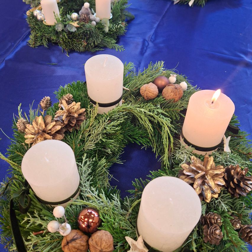 Ein Tisch geschmückt mit zwei Kränzen, einer mit vier brennenden Kerzen und Tannenzapfen und der andere mit einer einzelnen brennenden Kerze, alles vor einem blauen Hintergrund.
