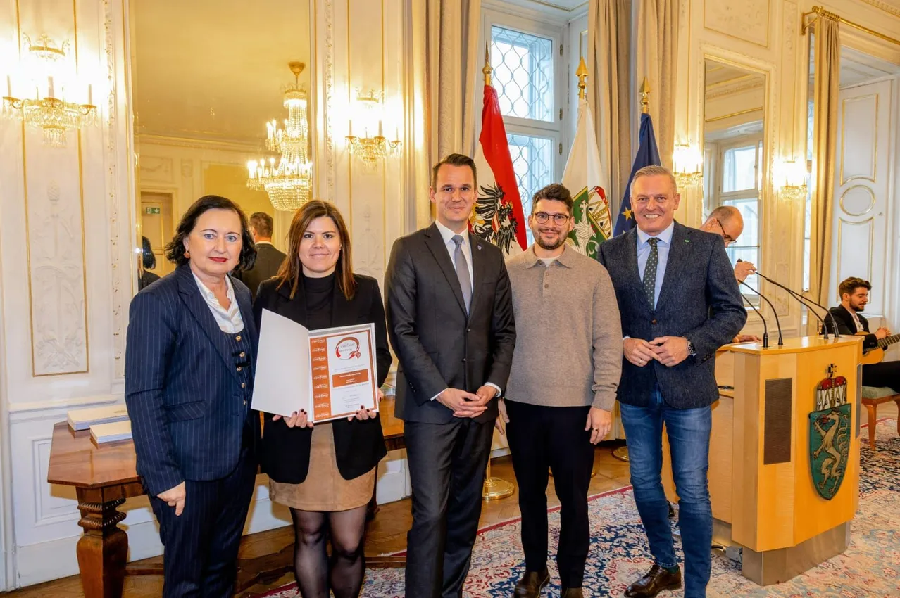 Fünf Personen stehen gemeinsam in einer formellen Umgebung, möglicherweise bei einer Preisverleihung. Eine Frau hält eine Preisurkunde. Hinter ihnen sind drei Flaggen sichtbar, und der Raum hat eine formelle Einrichtung.