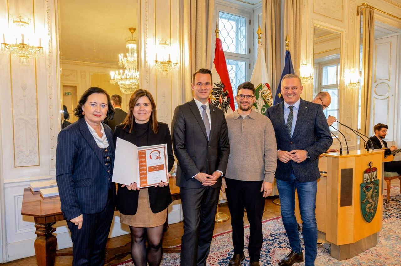 Fünf Personen stehen gemeinsam in einer formellen Umgebung, möglicherweise bei einer Preisverleihung. Eine Frau hält eine Preisurkunde. Hinter ihnen sind drei Flaggen sichtbar, und der Raum hat eine formelle Einrichtung.