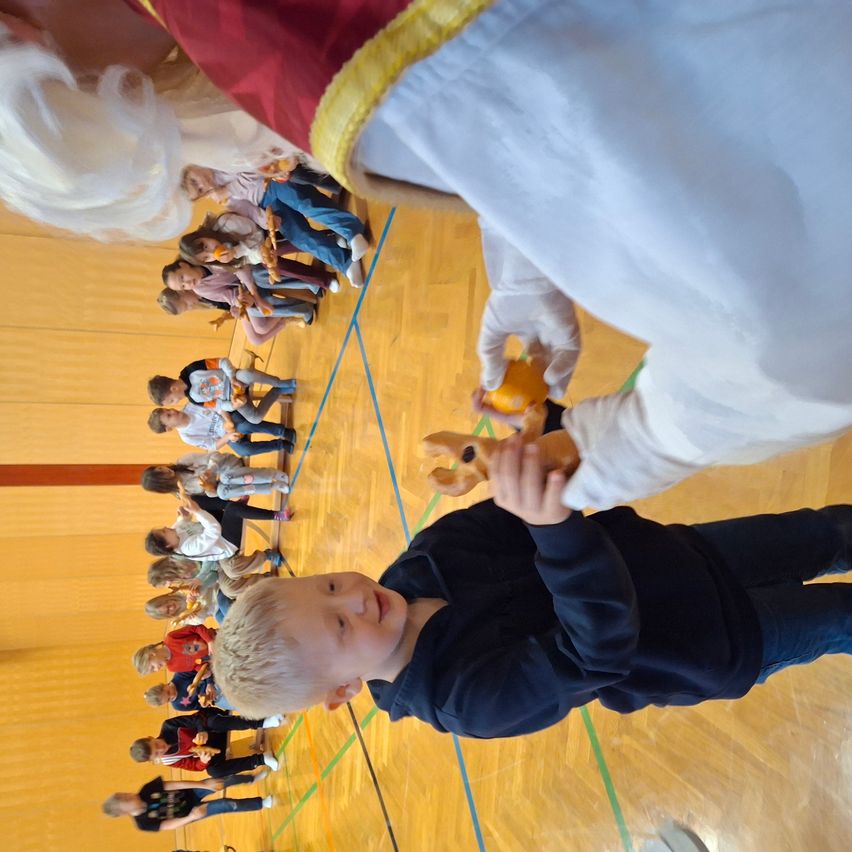 Ein junger Junge in einem blauen Shirt und Hosen erhält ein Spielzeug von einer Person in einem weißen und roten Gewand und weißen Handschuhen in einer Turnhalle.