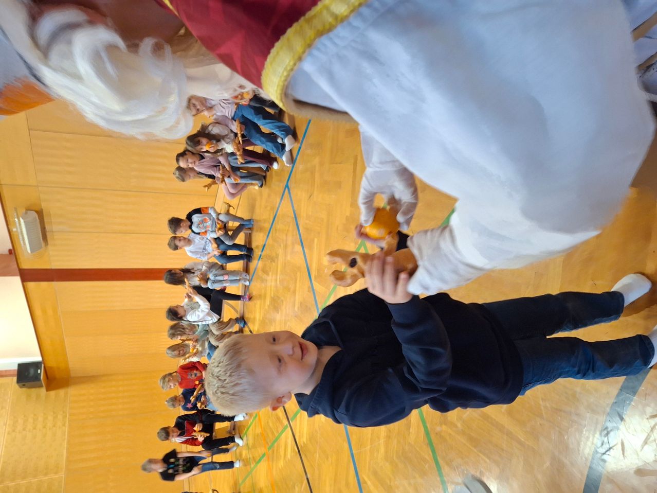 Ein junger Junge in einem blauen Shirt und Hosen erhält ein Spielzeug von einer Person in einem weißen und roten Gewand und weißen Handschuhen in einer Turnhalle.