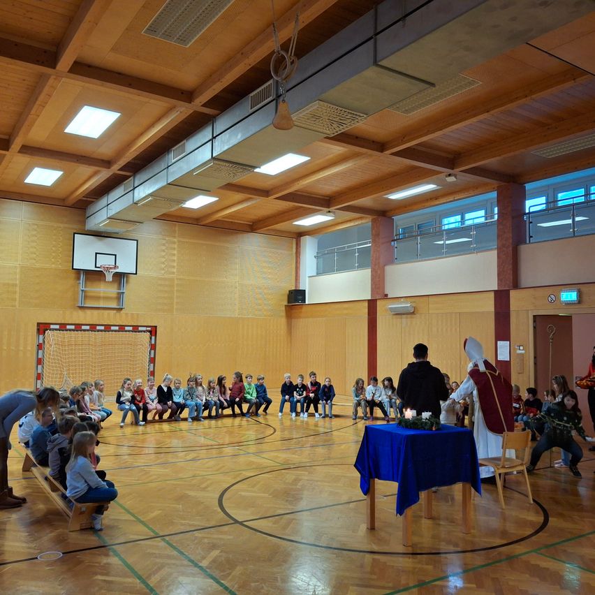 Eine Gruppe von Kindern sitzt auf einer Bank in einer Turnhalle. Eine Person steht auf einem Tisch mit einer Kerze. Ein Basketballkorb und Netz sind an der Wand montiert.