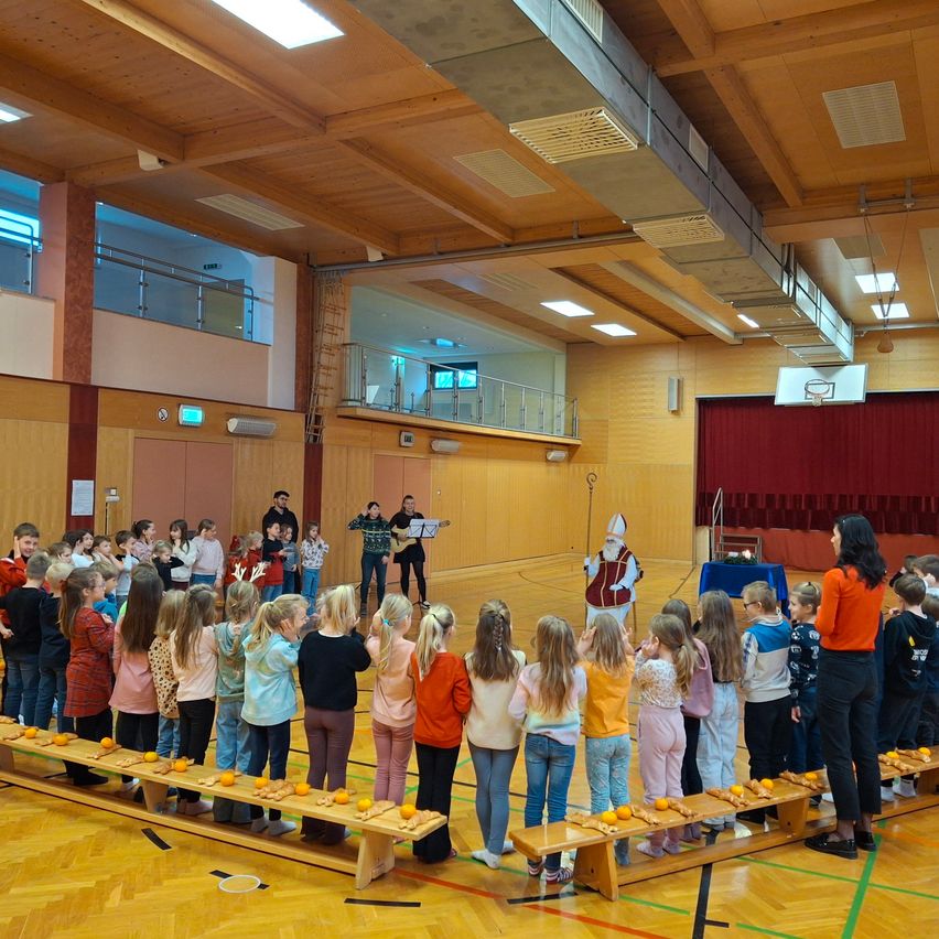 Eine Gruppe von Kindern und Erwachsenen steht in einem Saal. Sie stehen in einer Reihe mit Orangenscheiben auf einem Holztisch. Eine Person steht vor den Kindern und scheint zu singen. Ein Basketballkorb ist auf der rechten Seite. Eine Person steht auf der Bühne.