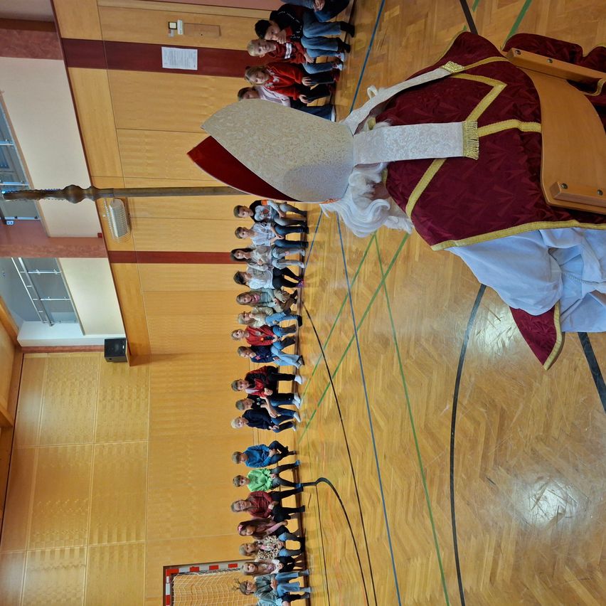Eine Gruppe von Kindern steht in einer Reihe und schaut auf eine als Bischof gekleidete Person in einer Turnhalle. Die Person hält einen Bischofsstab. Die Turnhalle hat Holzböden und -wände.
