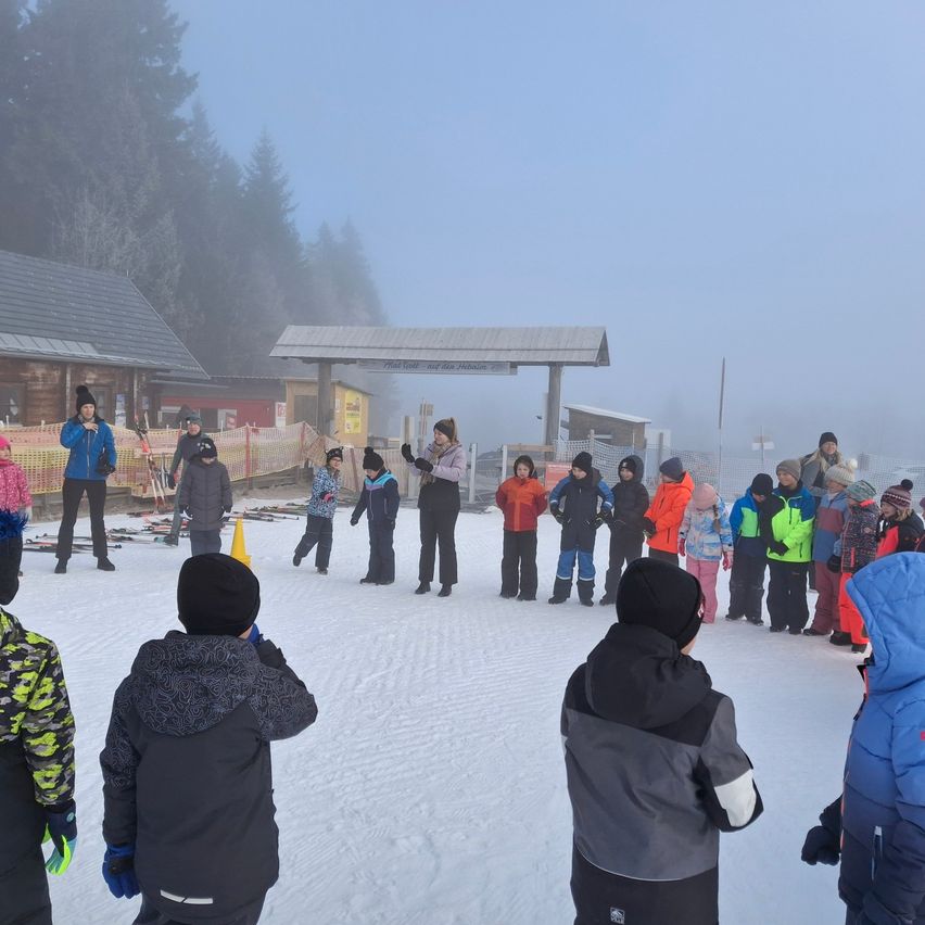 Eine Gruppe von Kindern und Erwachsenen in Winterkleidung steht auf einem verschneiten Feld. Sie sind von einem Zaun mit einem Schild umgeben. Bäume und ein Gebäude sind im Hintergrund.