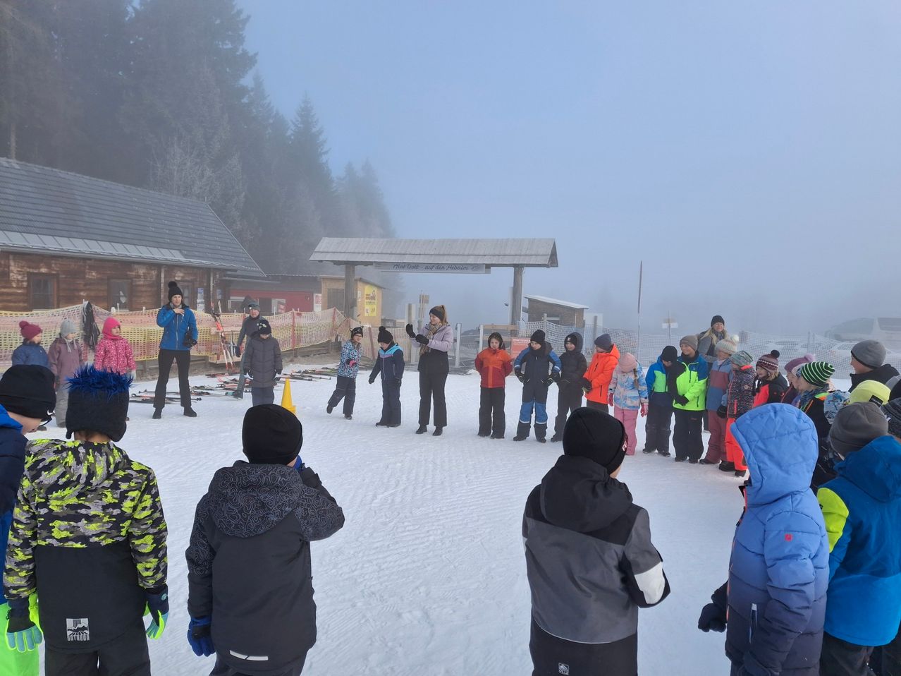 Eine Gruppe von Kindern und Erwachsenen in Winterkleidung steht auf einem verschneiten Feld. Sie sind von einem Zaun mit einem Schild umgeben. Bäume und ein Gebäude sind im Hintergrund.