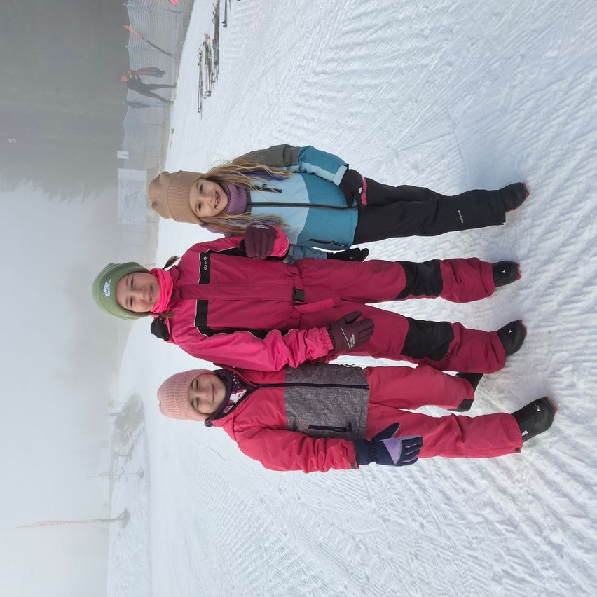 Drei Kinder stehen im Schnee. Das Kind vorne trägt einen rosa Mantel, während die anderen beiden blaue Jacken tragen. Sie alle lächeln für ein Foto.