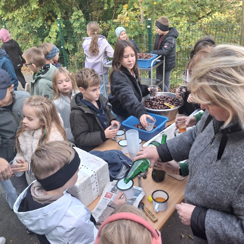 Eine Gruppe von Kindern und Erwachsenen versammelt sich um einen Tisch und bereitet Essen zu. Sie sind von verschiedenen Gegenständen wie Schüsseln, Bechern und Flaschen umgeben. Einige schauen auf das Essen, während andere sich auf ihre Aufgaben konzentrieren. Die Umgebung ist draußen mit Bäumen im Hintergrund.