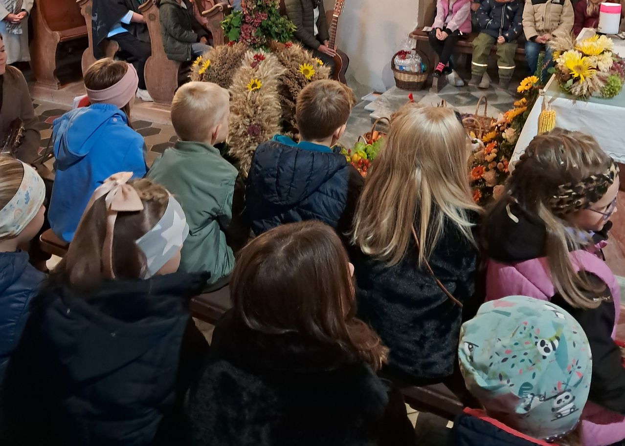 Kinder und Erwachsene versammeln sich in einer Kirche für eine Veranstaltung. Einige sitzen auf Bänken, während andere in der Nähe von Blumen und einer Gitarre stehen. Einige Erwachsene sitzen hinten.