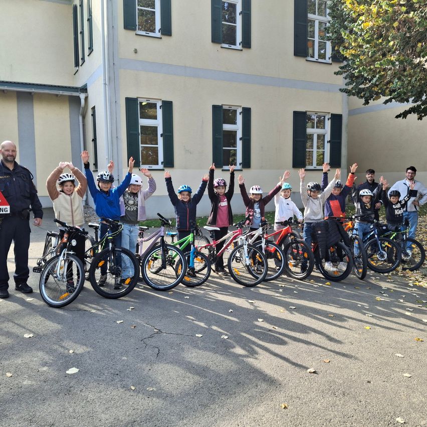 Eine Gruppe von Kindern in Helmen posiert mit ihren Fahrrädern vor einem Gebäude mit einem Mann und einem Polizisten.