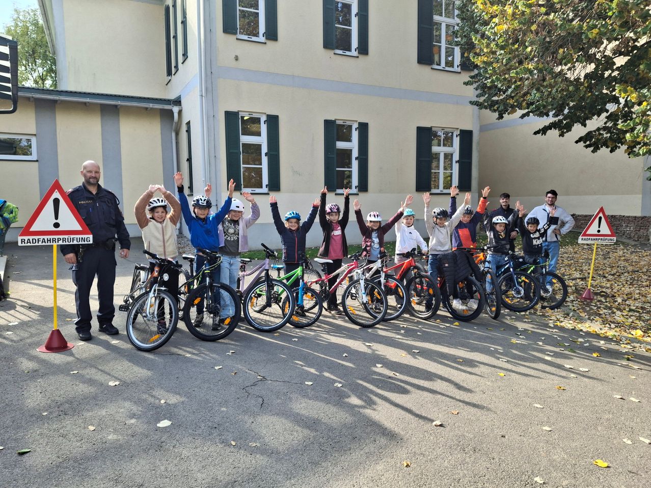 Eine Gruppe von Kindern in Helmen posiert mit ihren Fahrrädern vor einem Gebäude mit einem Mann und einem Polizisten.