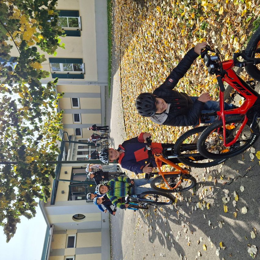 Mehrere Kinder posieren mit ihren Fahrrädern auf einem Pfad mit Laub im Herbst. Es gibt ein Gebäude mit Glasfenstern und einen Baum mit grünen Blättern.