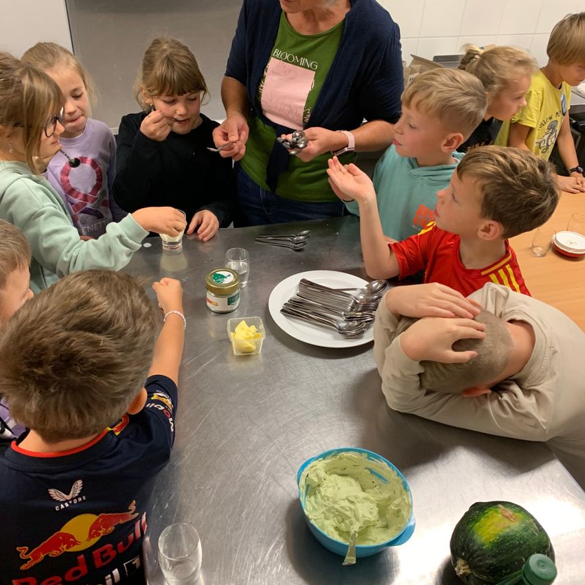 Eine Gruppe von Kindern sitzt um einen Tisch in einer Küche herum. Sie schauen auf eine Frau, die einen Löffel hält und ihnen etwas zeigt. Auf dem Tisch sind Teller, Löffel und eine Schüssel mit Essen. Die Kinder tragen verschiedenfarbige Hemden.