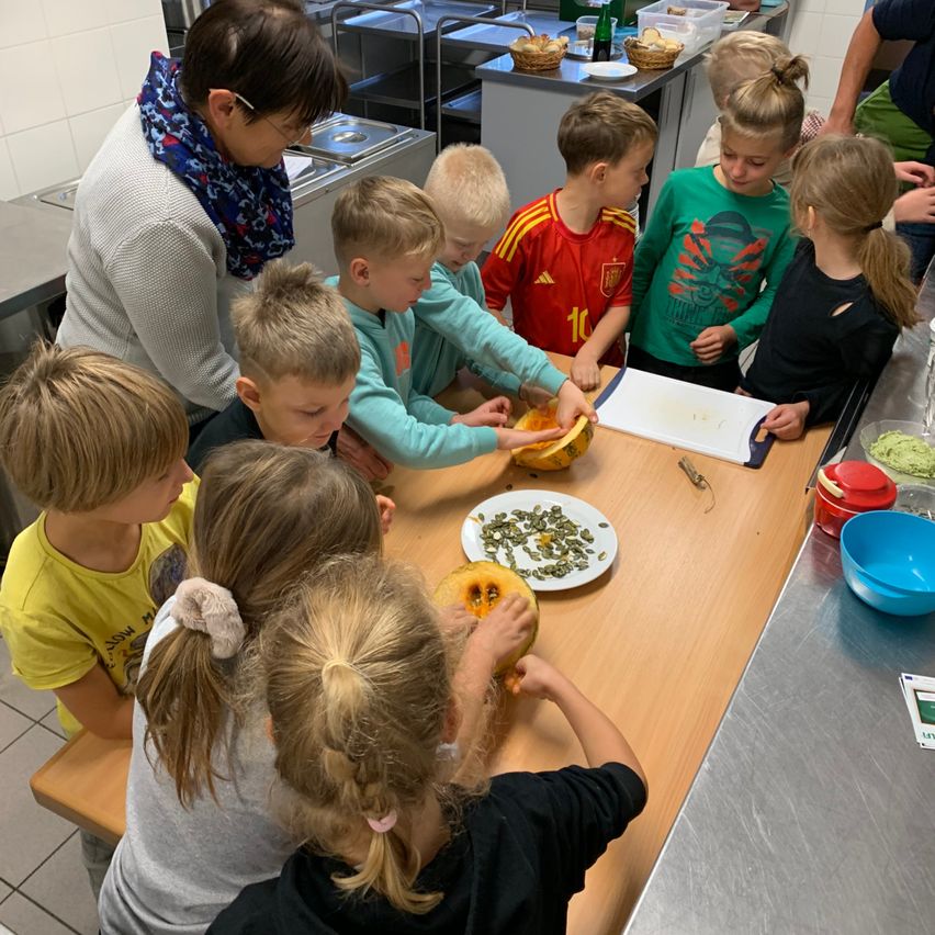 Eine Gruppe von Kindern und einem Erwachsenen versammelt sich um einen Tisch und schnitzelt Kürbisse. Der Tisch hat einen Teller mit Samen, eine Schüssel und ein Schneidebrett. Der Raum hat Edelstahl-Theken und Regale.