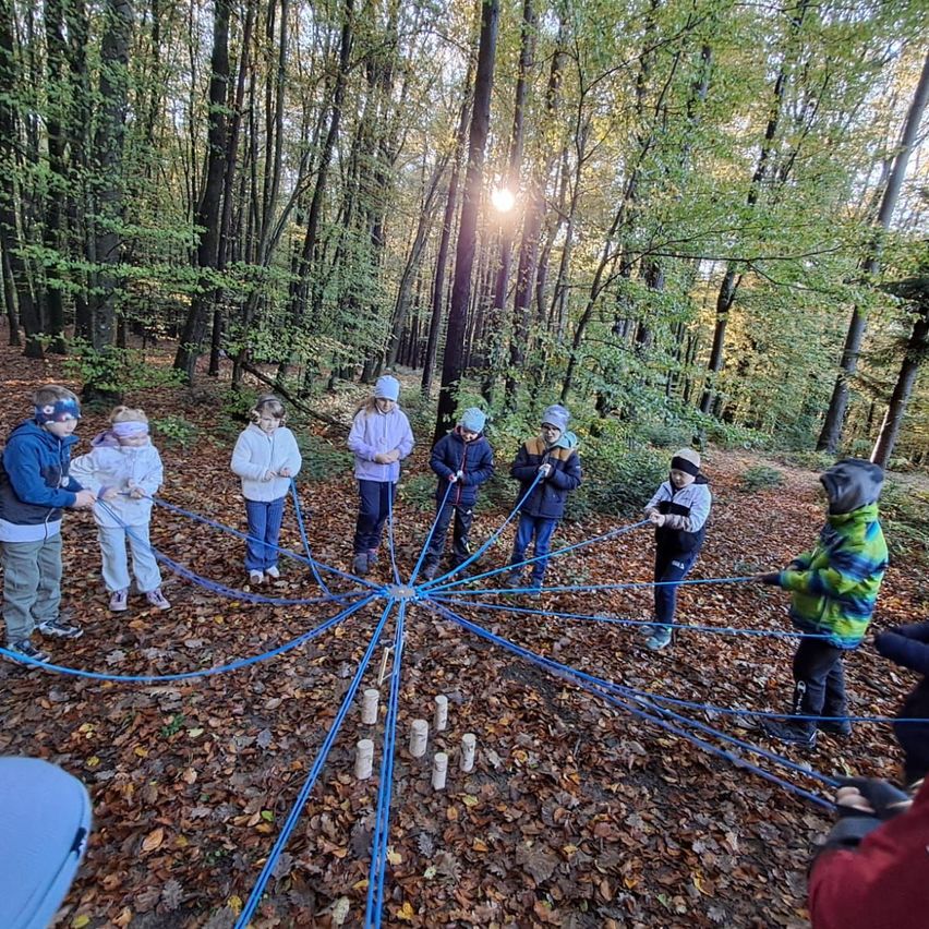 Eine Gruppe von Kindern steht in einem Kreis im Wald, hält blaue Seile und spielt ein Spiel.