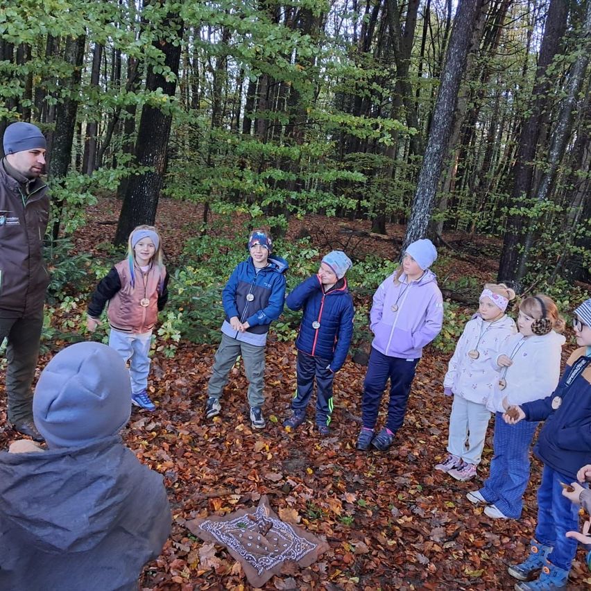 Eine Gruppe von Kindern und einem Erwachsenen steht im Wald. Der Erwachsene steht vor den Kindern und scheint Anweisungen zu geben.