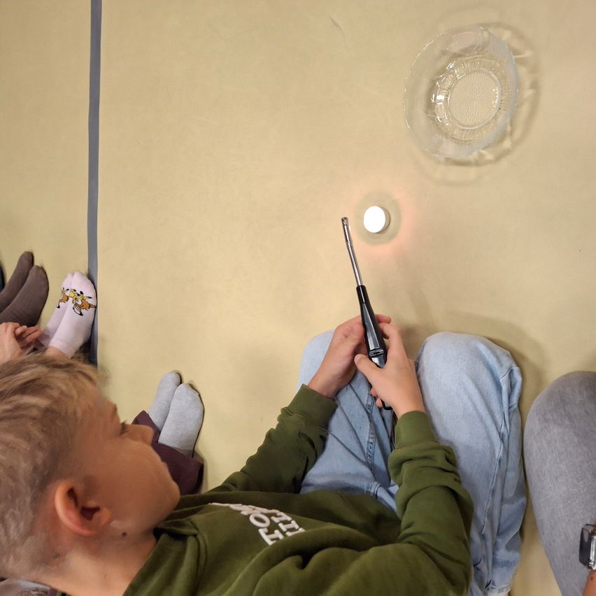 Ein junger Junge sitzt auf dem Boden und hält einen Schraubendreher in der Hand. Er untersucht ein kleines weißes Objekt. Ihn umgeben andere, und eine Platte hängt von der Decke.