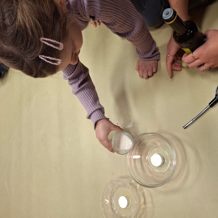 Ein Kind mischt eine Substanz in einer Schüssel auf einem Tisch, während eine andere Person eine Flasche Flüssigkeit hält. Es gibt zwei Schüsseln mit Kerzen und eine Schere in der Nähe.