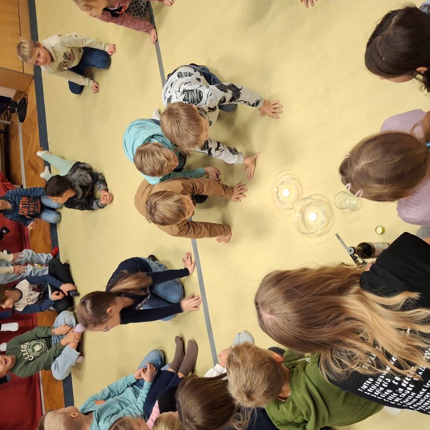 Eine Gruppe von Kindern versammelt sich im Kreis, einige knien auf einem gelben Boden und nehmen an einer Klassenaktivität teil. Einige Kerzen sind angezündet und in der Mitte platziert.