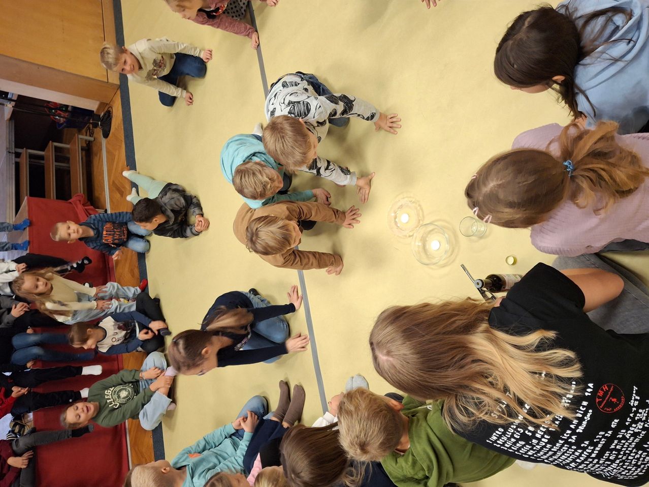 Eine Gruppe von Kindern versammelt sich im Kreis, einige knien auf einem gelben Boden und nehmen an einer Klassenaktivität teil. Einige Kerzen sind angezündet und in der Mitte platziert.