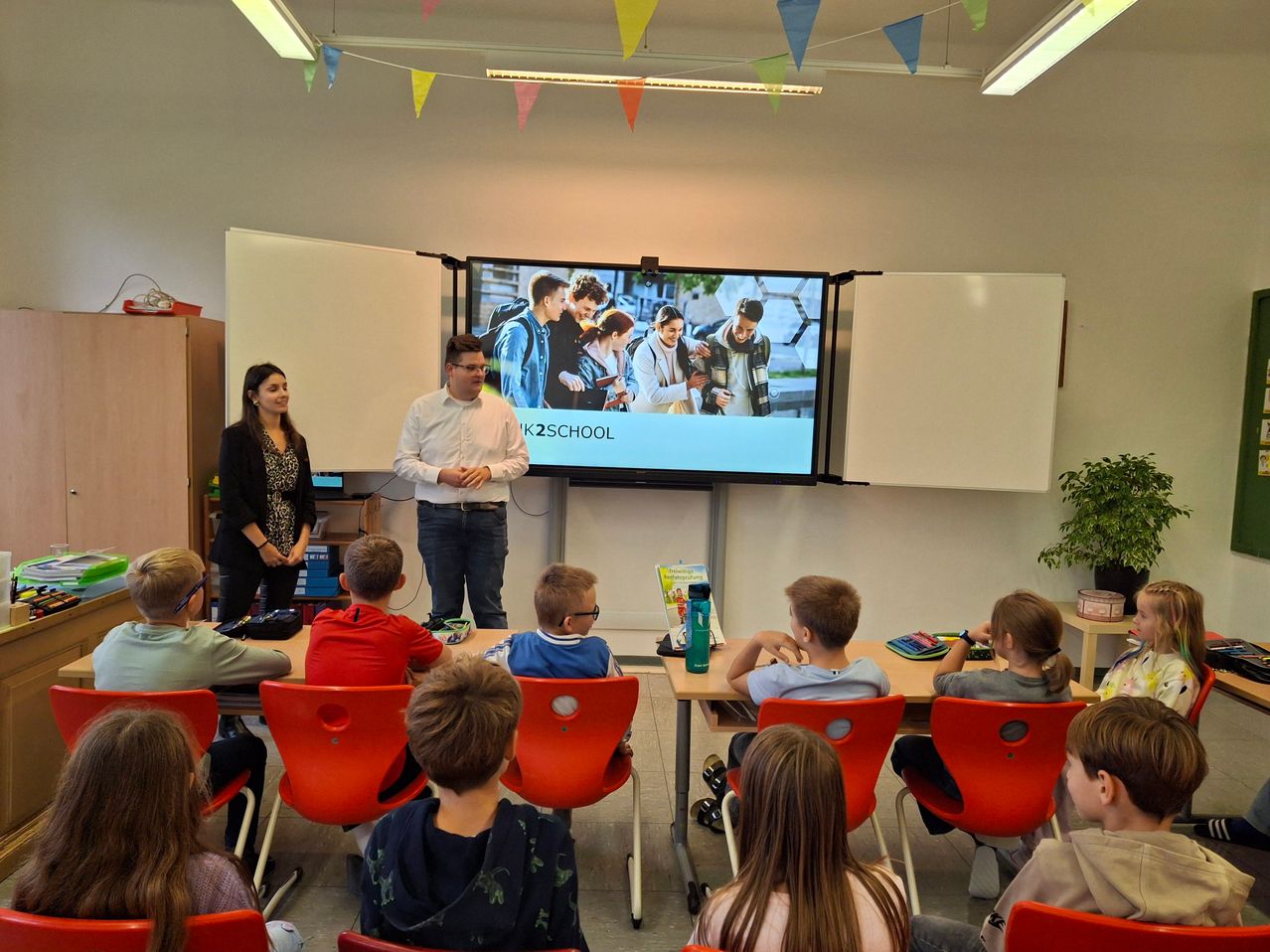 Ein Klassenzimmer mit einem Mann und einer Frau, die vor einer Projektionsleinwand stehen, auf der eine Gruppe von Schülern zu sehen ist. Die Schüler sitzen in roten Stühlen und beobachten aufmerksam die Präsentation.