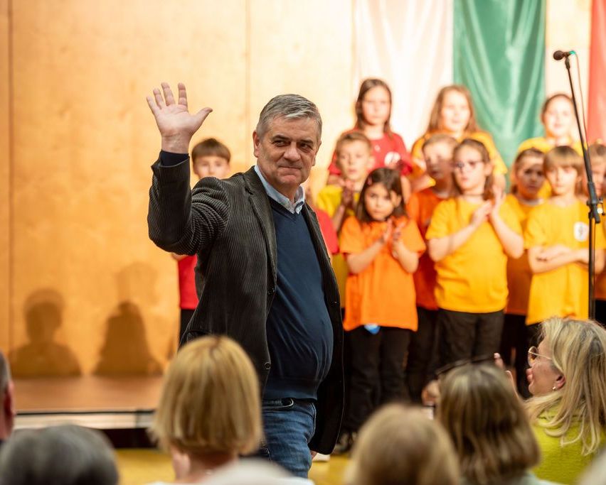 Ein Mann winkt einer Gruppe von Kindern in gelben Shirts auf der Bühne zu. Hinter ihm ist ein grüner Banner zu sehen. Das Publikum sitzt vor ihm und schaut zu.