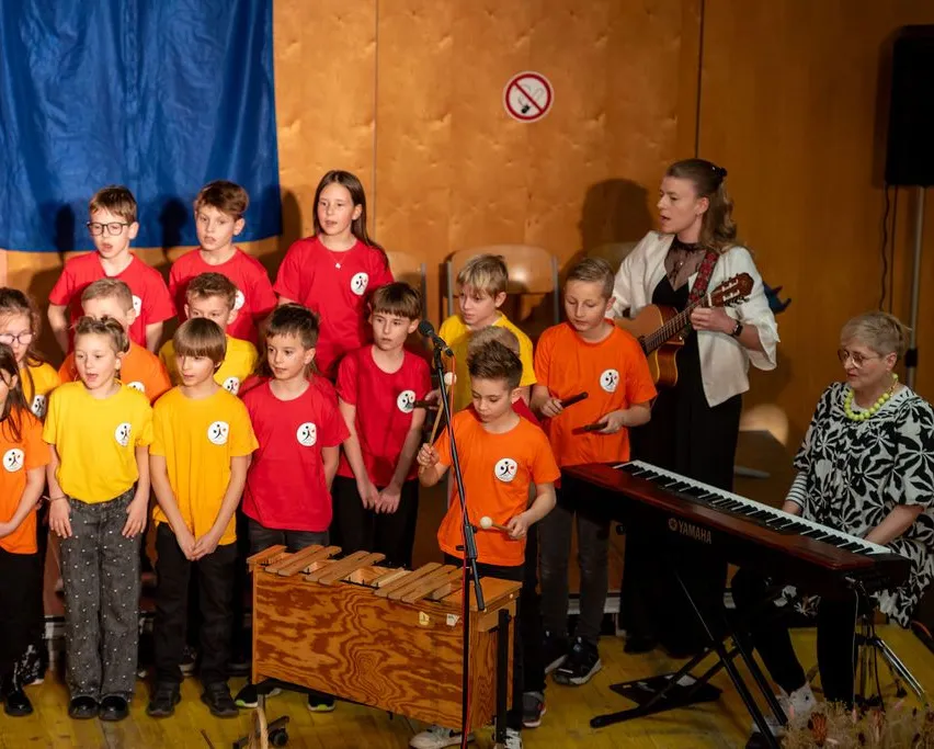 Eine Gruppe von Kindern in roten und gelben T-Shirts steht und hält Instrumente, wahrscheinlich bei einer Aufführung in einer Schule. Eine Frau steht hinter ihnen, hält eine Gitarre und spielt Klavier.