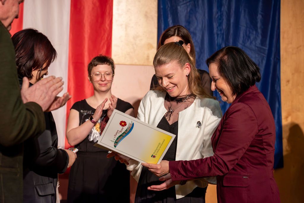 Eine Frau erhält einen Preis und lächelt, während andere applaudieren. Die Frau hält eine Urkunde mit Logo und Text. Eine Frau in einem roten Blazer reicht ihr die Urkunde. Im Hintergrund sind eine Flagge und ein blauer Vorhang zu sehen.