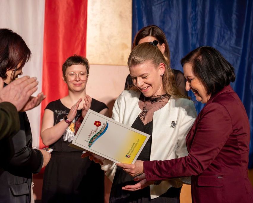 Eine Frau erhält einen Preis und lächelt, während andere applaudieren. Die Frau hält eine Urkunde mit Logo und Text. Eine Frau in einem roten Blazer reicht ihr die Urkunde. Im Hintergrund sind eine Flagge und ein blauer Vorhang zu sehen.