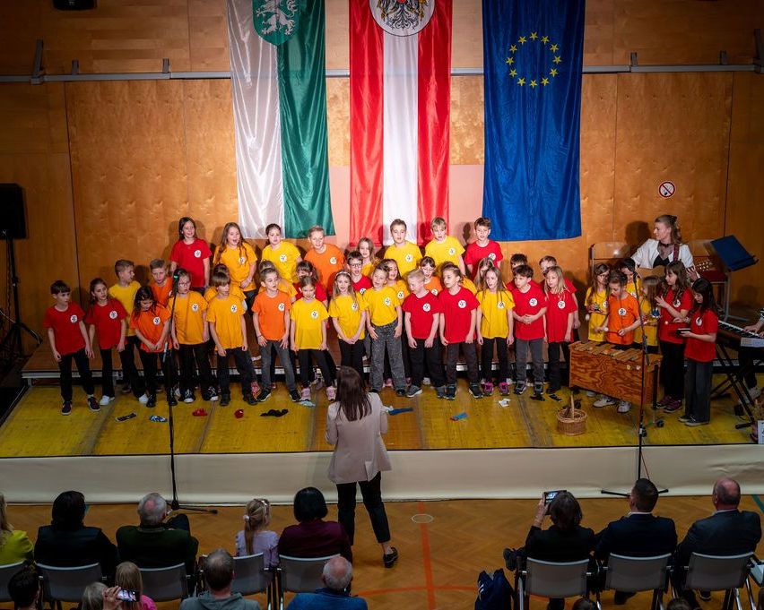 Eine Gruppe von Kindern in einheitlichen Shirts tritt mit Musikinstrumenten auf der Bühne auf. Dahinter befinden sich Flaggen von Ungarn, Österreich und der EU. Ein Publikum sitzt davor.