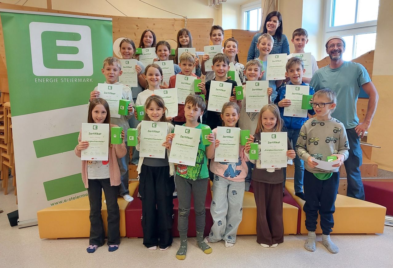 Eine Gruppe von Kindern, einige in Winterkleidung, halten Zertifikate und grüne Becher vor einem Banner. Eine Frau lächelt im Hintergrund.