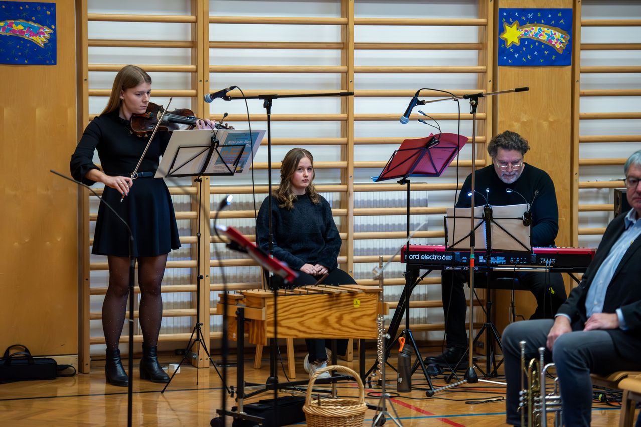 Drei Musiker spielen in einem Raum. Eine Geigerin steht links, ein Pianist mit einem Keyboard sitzt rechts und ein Perkussionist mit einem Xylophon sitzt in der Mitte. Alle haben Notenständer vor sich.