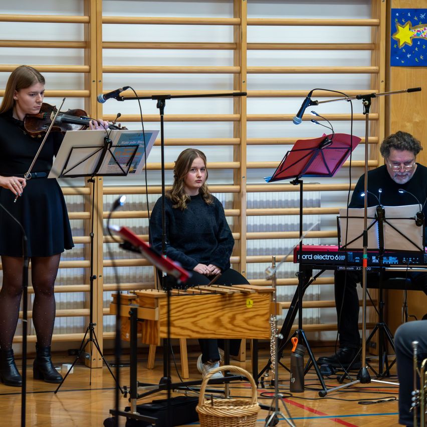 Drei Musiker spielen in einem Raum. Eine Geigerin steht links, ein Pianist mit einem Keyboard sitzt rechts und ein Perkussionist mit einem Xylophon sitzt in der Mitte. Alle haben Notenständer vor sich.