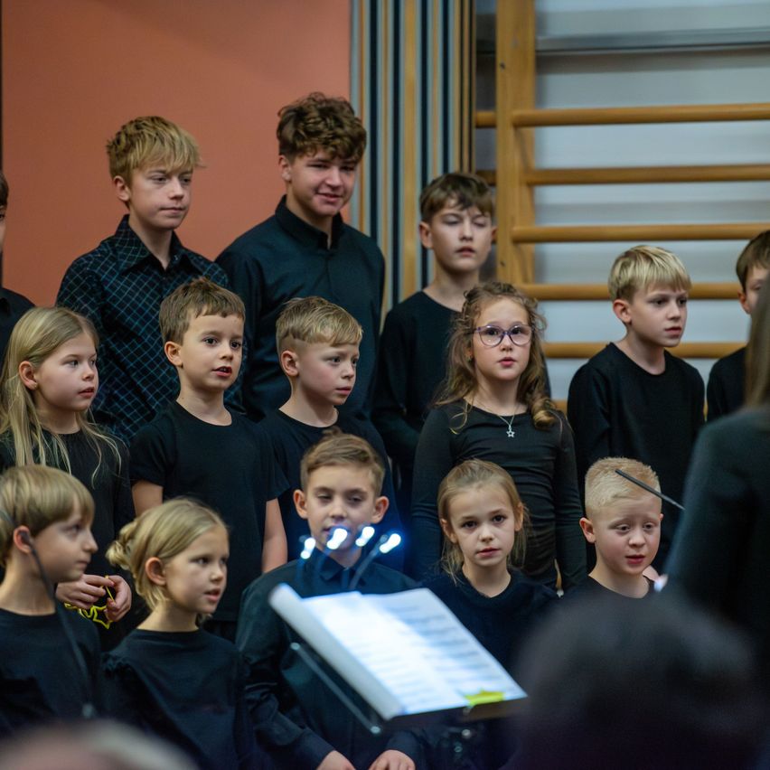 Ein Chor von Kindern, Jungen und Mädchen, gekleidet in Schwarz, tritt auf einer Bühne mit einem Hintergrund aus warmen, pfirsichfarbenen Wänden auf. Sie wirken konzentriert, mit einem Dirigenten vor ihnen.