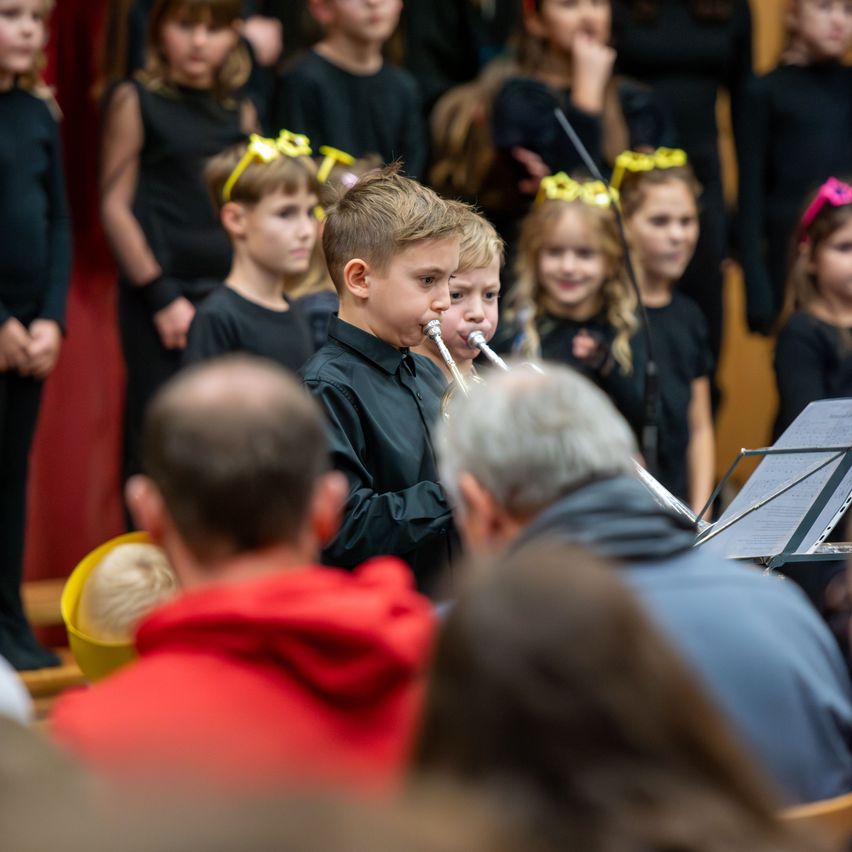 Kinder in schwarzen Outfits stehen in einem Chor und spielen Trompeten. Ein Junge in der Mitte konzentriert sich auf sein Instrument. Dahinter beobachtet eine verschwommene Figur Musikblätter. Im Vordergrund sieht ein Mann aufmerksam zu.