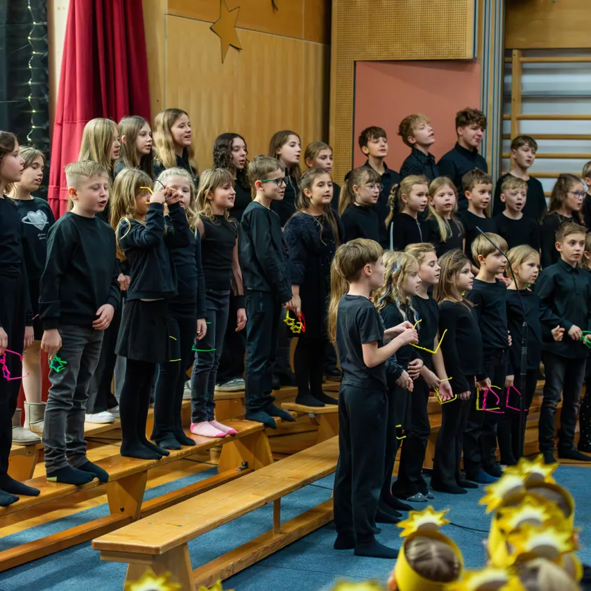 Ein Chor von Kindern, in Schwarz gekleidet, singt in einem Klassenzimmer mit Holzbänken, einige tragen gelbe Stirnbänder mit Sternen.